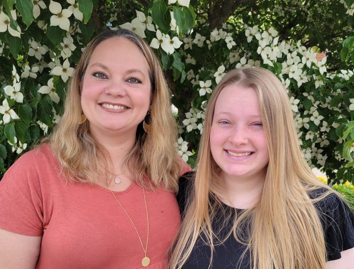 Deana Cummings stands with Heather smiling in front of blooming spring flowers.