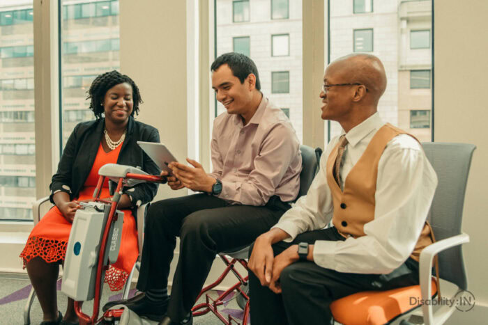 three people, diverse in ability and background, sit in an office waiting room talking.