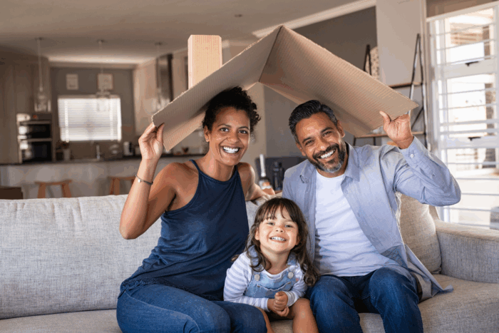 Smiling mother, father, daughter sit on couch holding house roof over the family.
