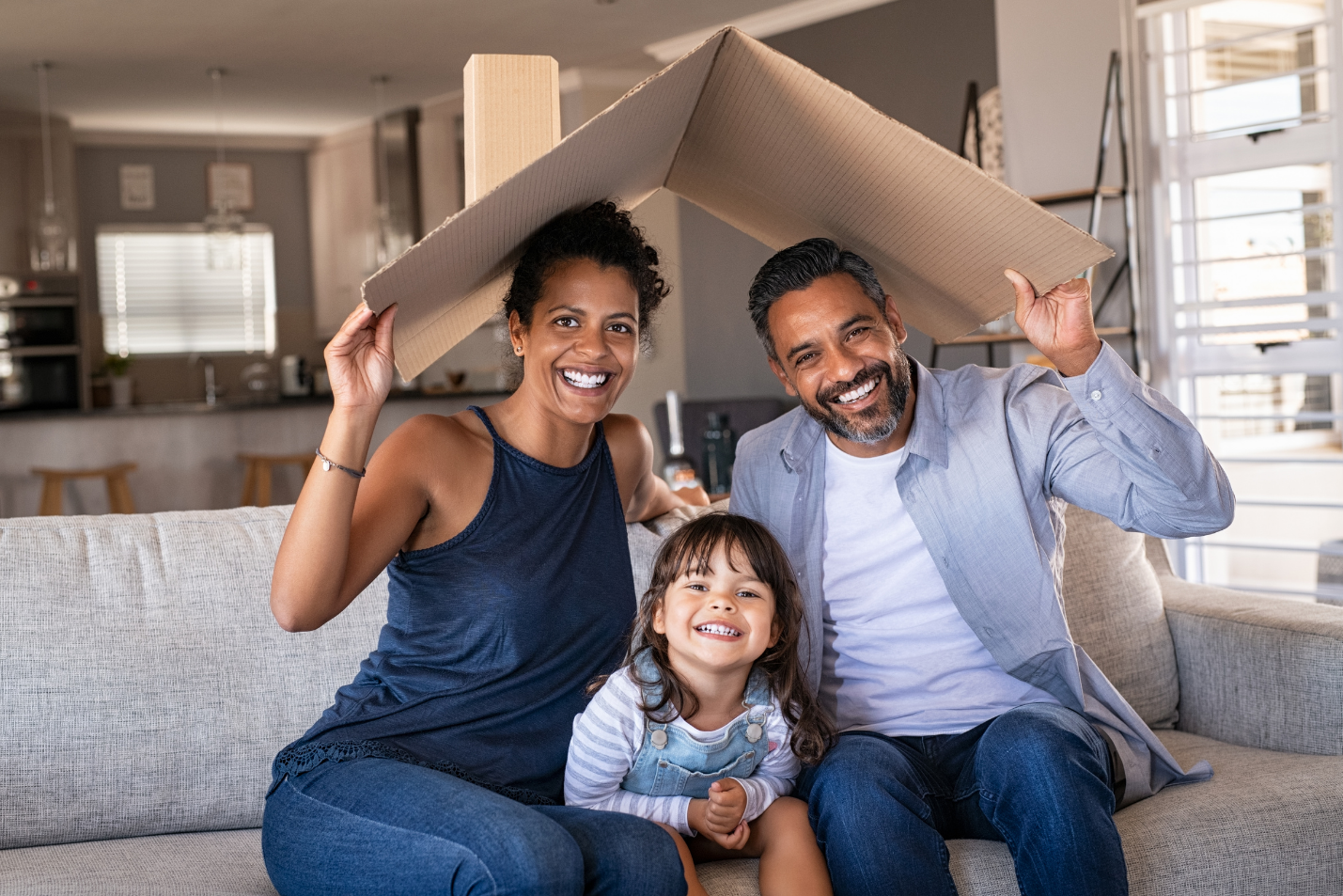 Smiling mother, father, daughter sit on couch holding house roof over the family.