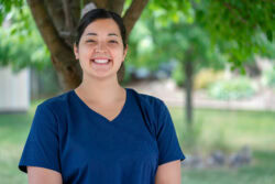 Nurse in scrubs smiles at camera while she stands outside.